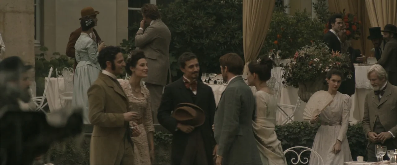 Eiffel (2021) movie still: fullbody — A group of people in formal turn-of-the-century attire socialize at an outdoor garden party. The guests stand…