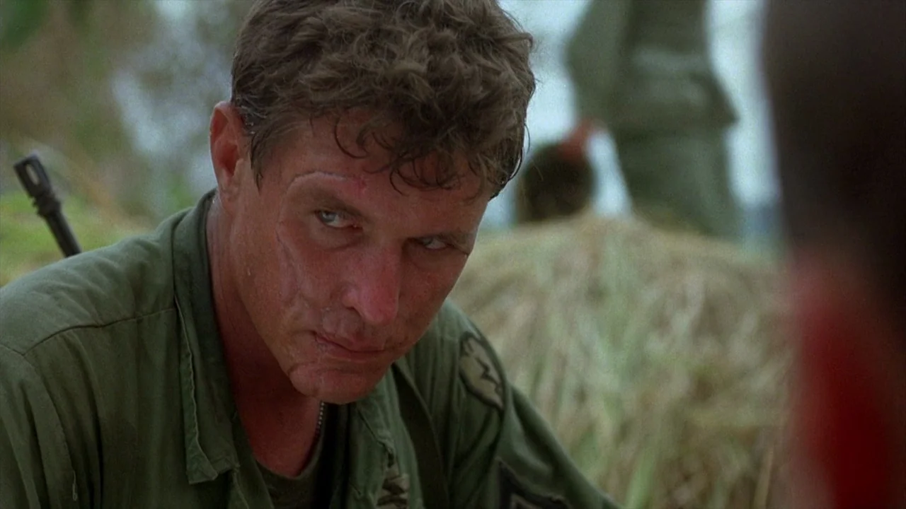 Platoon (1986) movie still: medium — An outdoor location with blurred foliage and a haystack in the background. A soldier with a visible facial scar…