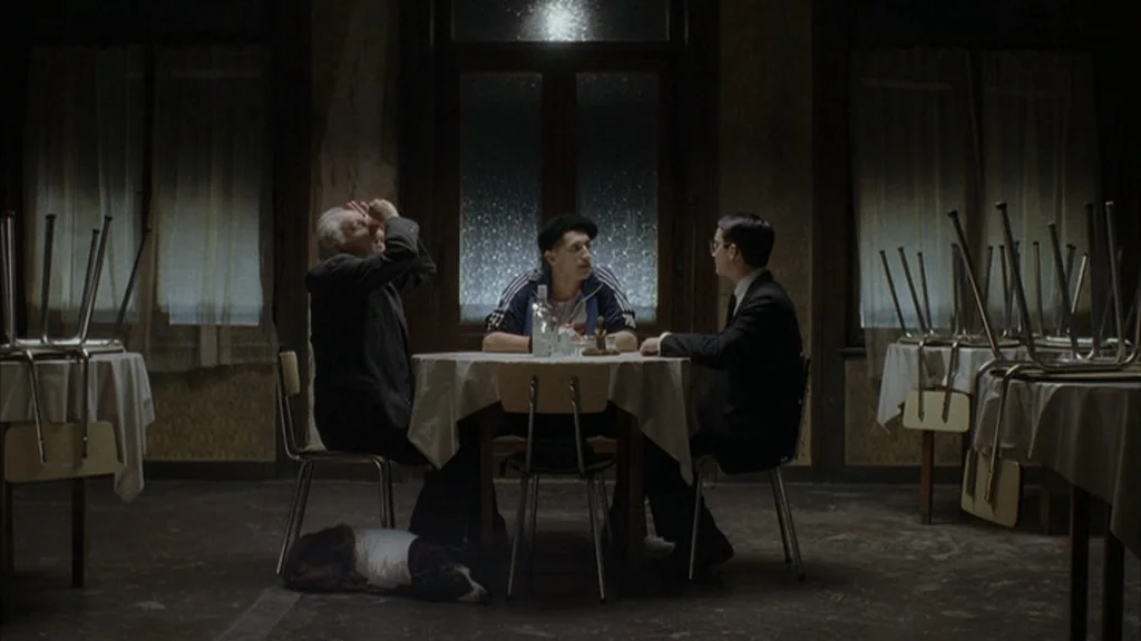 Everything Is Illuminated (2005) movie still: wide — A dimly lit cafe features tables with chairs stacked upside down on them. Three men sit around…