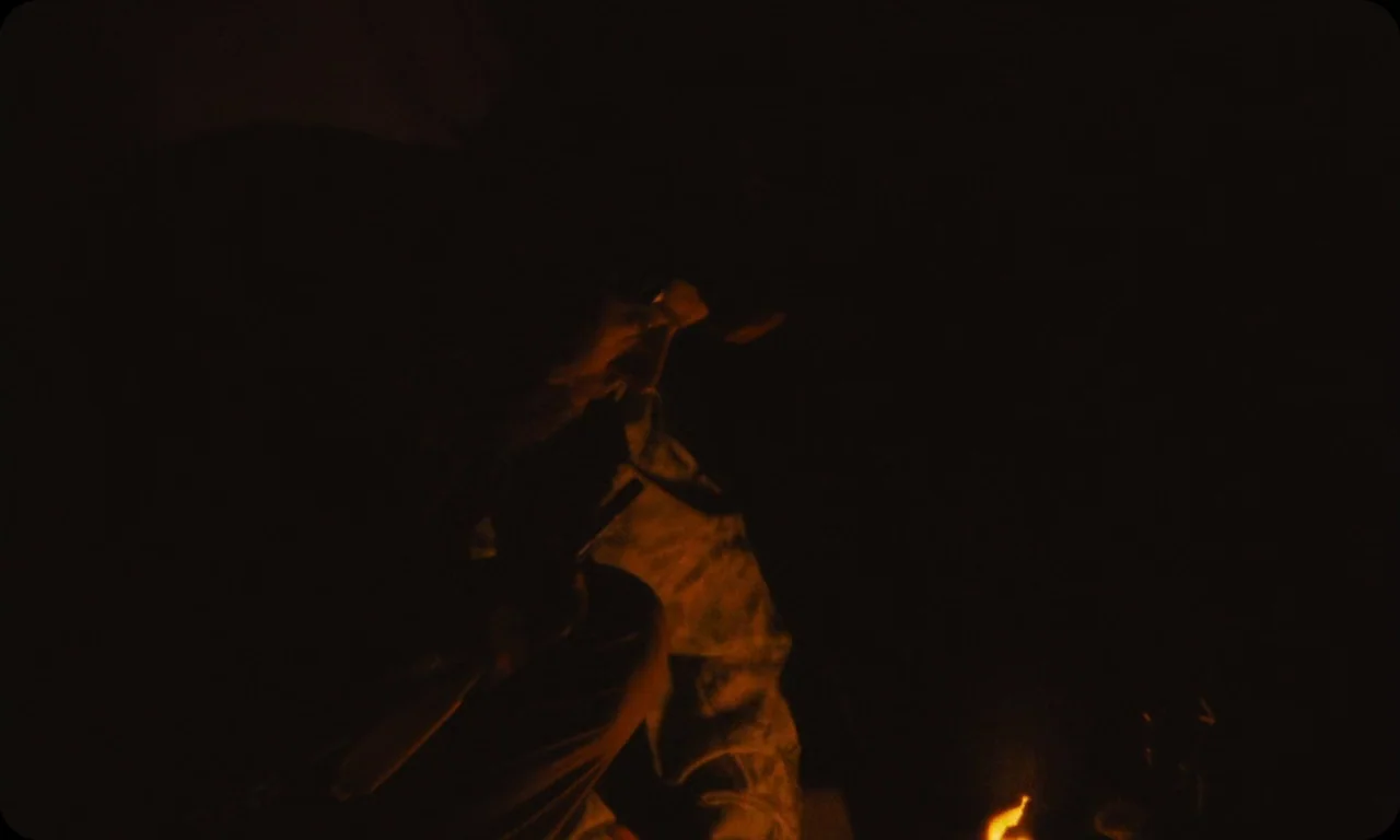 Vulcanizadora (2024) movie still: medium — An extremely dark scene features a person crouching in a campsite at night. The man holds an object while…