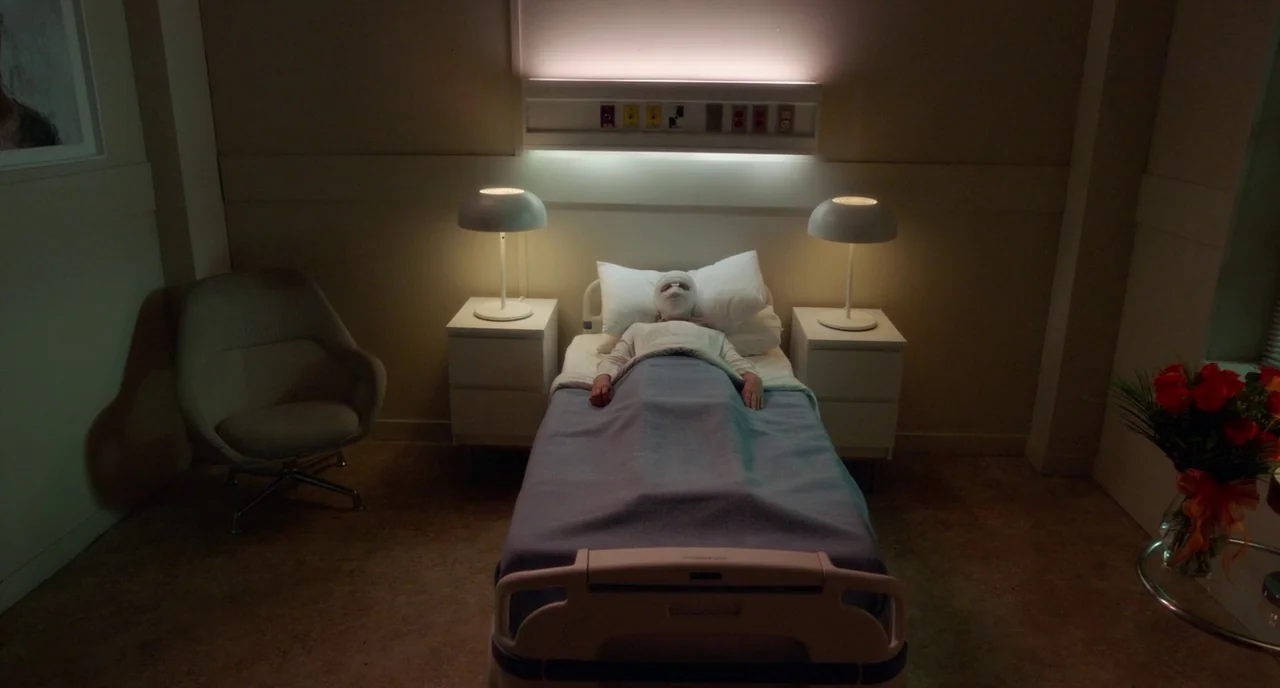 Rabid (2019) movie still: wide — A quiet hospital room with a patient centered in a narrow bed. Minimalist furniture surrounds the bed with a small…