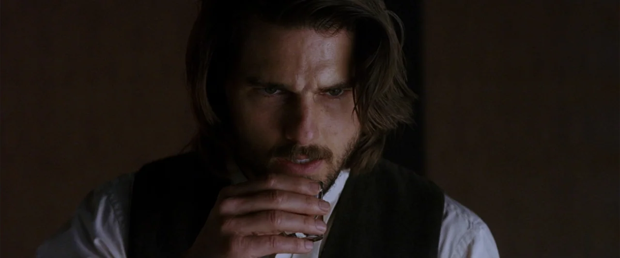 The Last Samurai (2003) movie still: closeup — A dimly lit interior space with a dark, indistinct background. A man with shoulder-length hair holds…