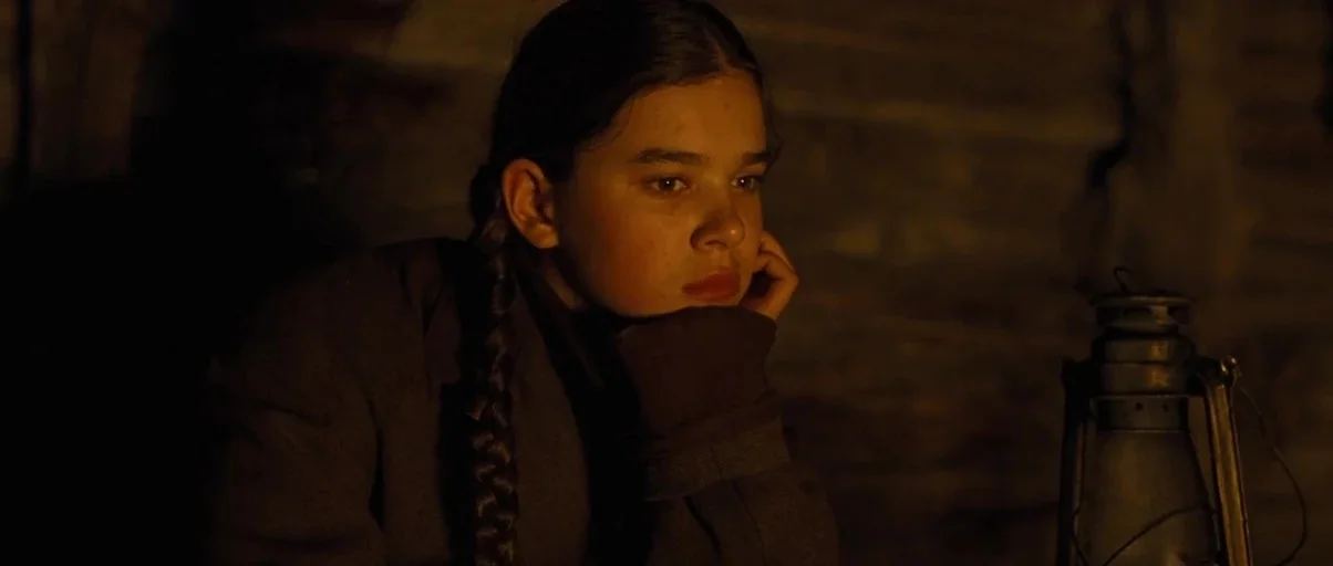 True Grit (2010) movie still: closeup — A dark wooden room with logs visible in the background wall. A young girl rests her chin on her hand while…