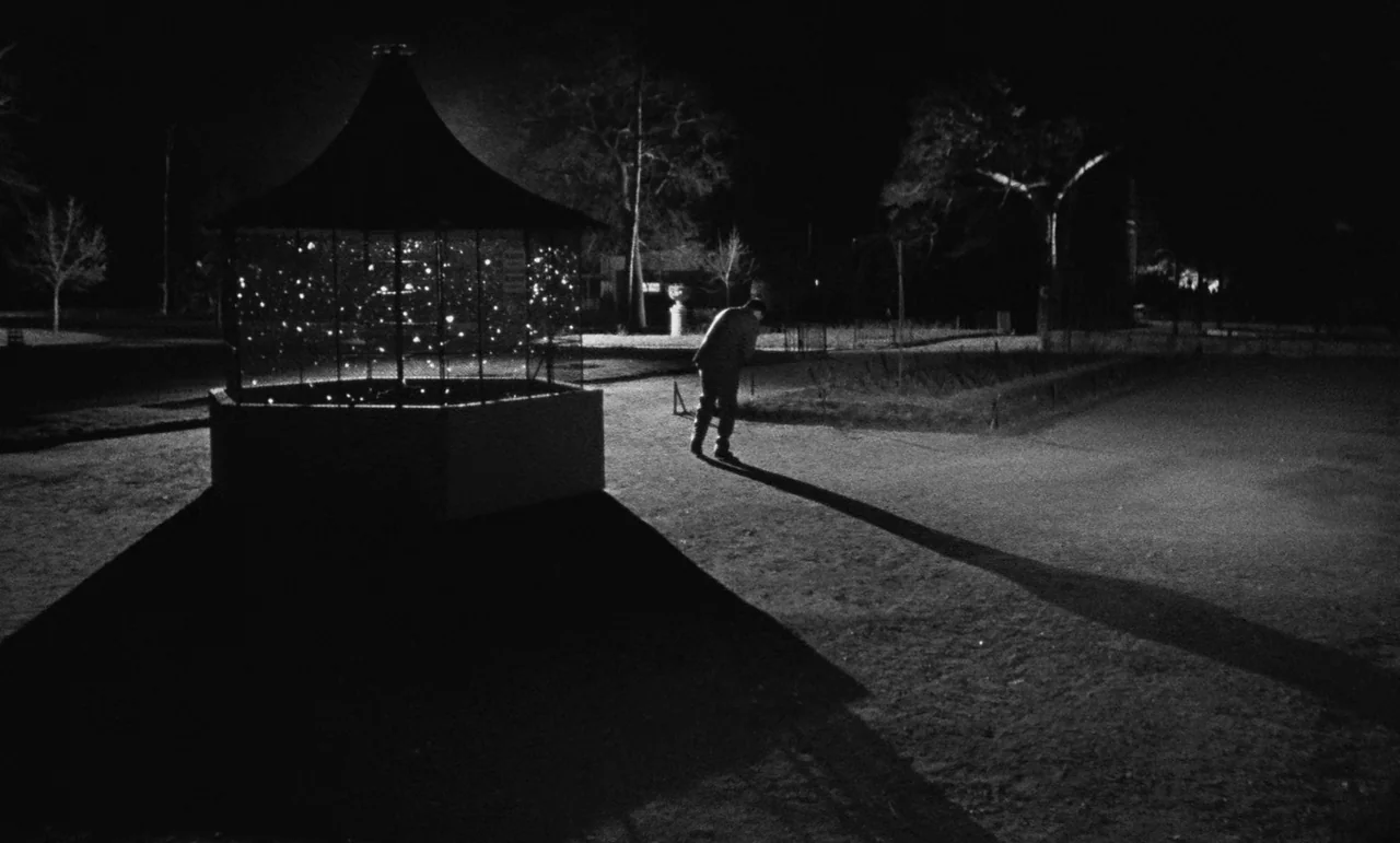Witness In The City (1959) movie still: wide — A night scene in a park featuring a illuminated gazebo casting long sharp shadows. A solitary person…