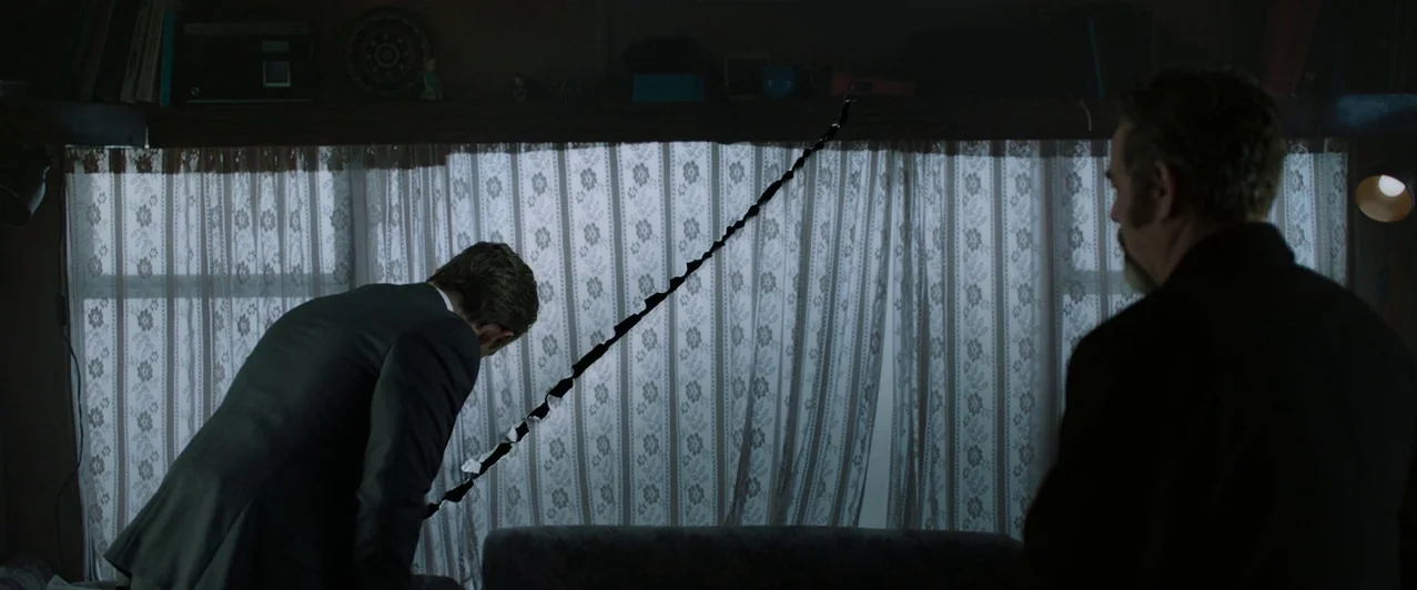 Ghost Stories (2017) movie still: wide — A cluttered, dimly lit camper interior features floral lace curtains over a small window. One man stands by…