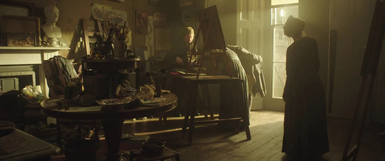 Mr. Turner (2014) movie still: wide — A cluttered 19th-century artist studio contains easels, a table with brushes, and wall art. People stand in…