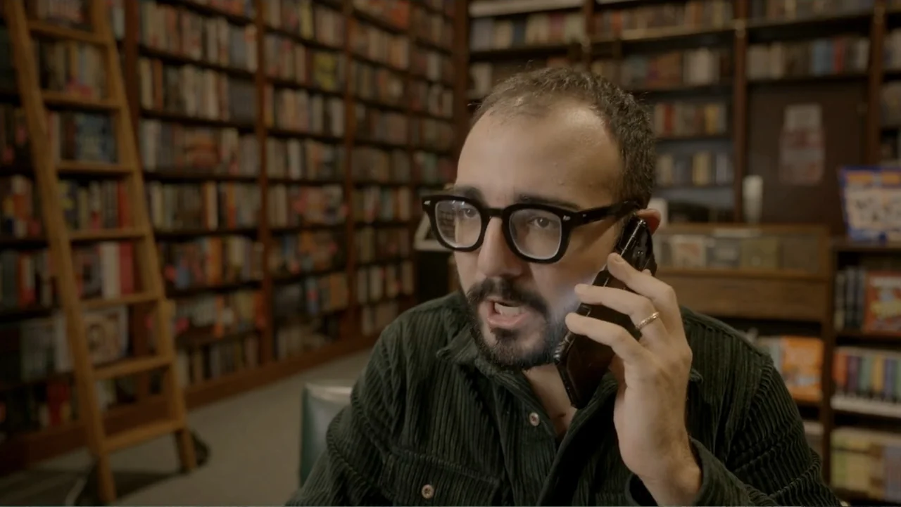 Making of: Scars of Beauty (2025) movie still: medium — A man stands in front of tall wooden shelves packed with books. He holds a smartphone to his…