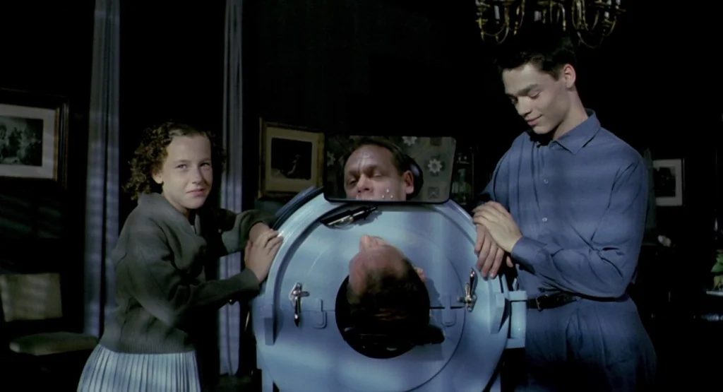 In a Glass Cage (1986) movie still: medium — A dark, dimly lit room contains an iron lung occupying the center space. Two young people stand on…