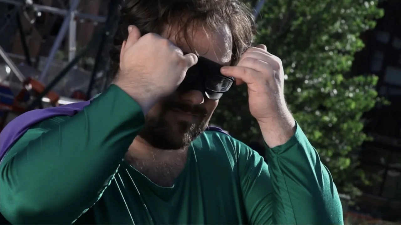 Hello I Love You (2018) movie still: closeup — A man in a green superhero costume stands outdoors in front of a structure. He is using both hands to…