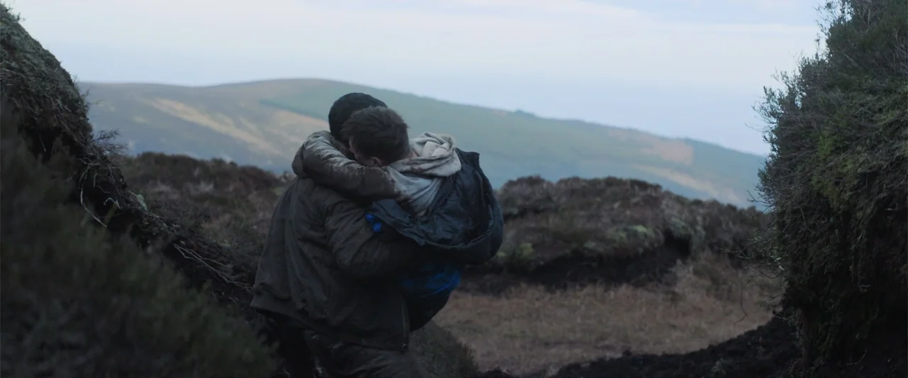 Bring Them Down (2025) movie still: medium — A desolate hillside covered in low vegetation and earth under an overcast sky. One man carries an…