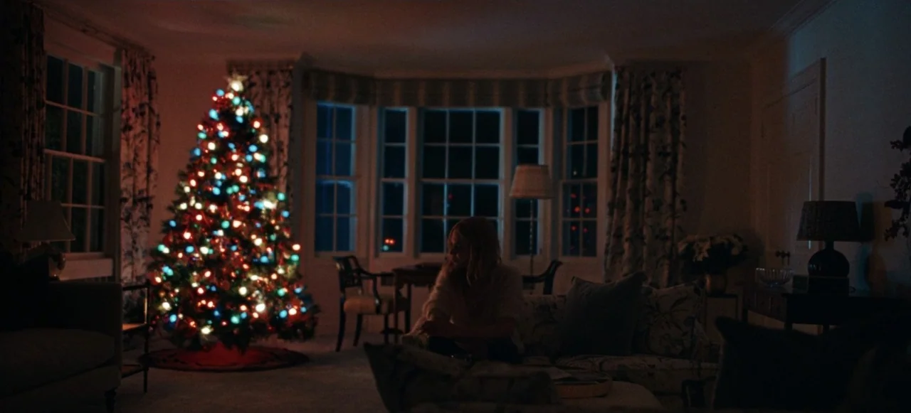Babygirl (2024) movie still: wide — A dimly lit room at night features a large, glowing, multi-colored Christmas tree in the corner. A woman sits on…