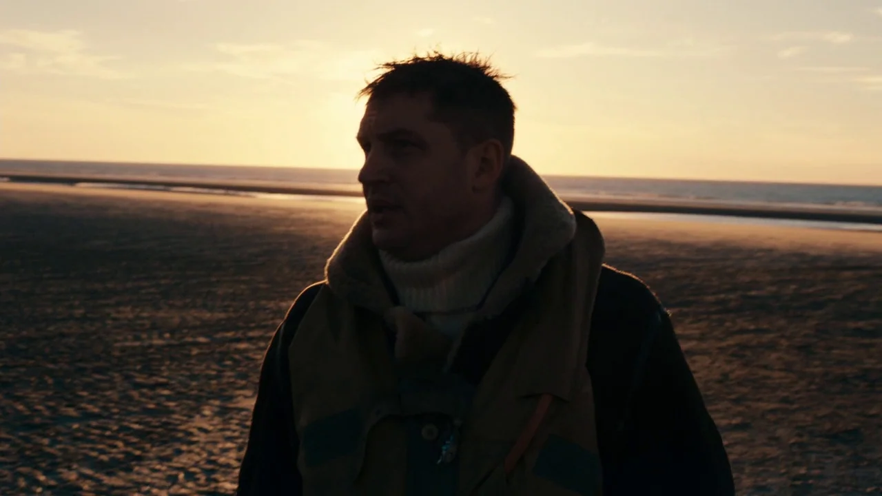 Dunkirk (2017) movie still: medium — A vast, empty coastline at sunset with calm water meeting dark, wet sand. A man stands in the foreground, his…