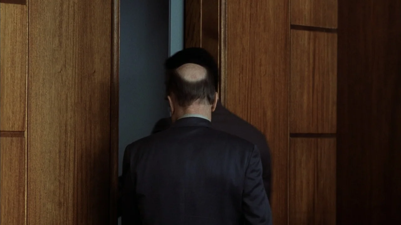 John Q (2002) movie still: medium — A man in a dark suit stands before a set of wood-paneled doors. He is positioned in front of an open elevator car.