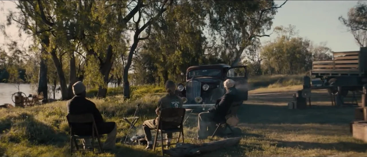 Fear Below (2025) movie still: wide — A rural campsite near water features tall trees and a vintage automobile parked nearby. Three people sit on…