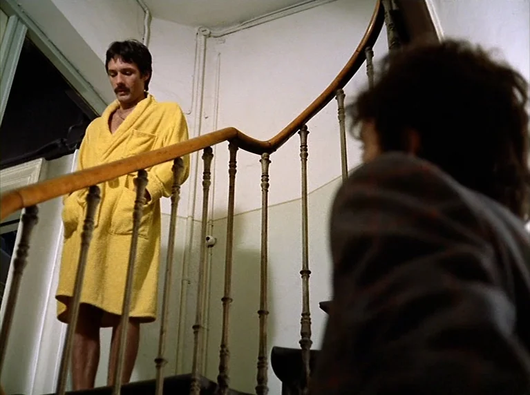 Fox and His Friends (1975) movie still: medium — A man in a yellow robe stands on a wooden staircase with iron railings. He looks down at a person…