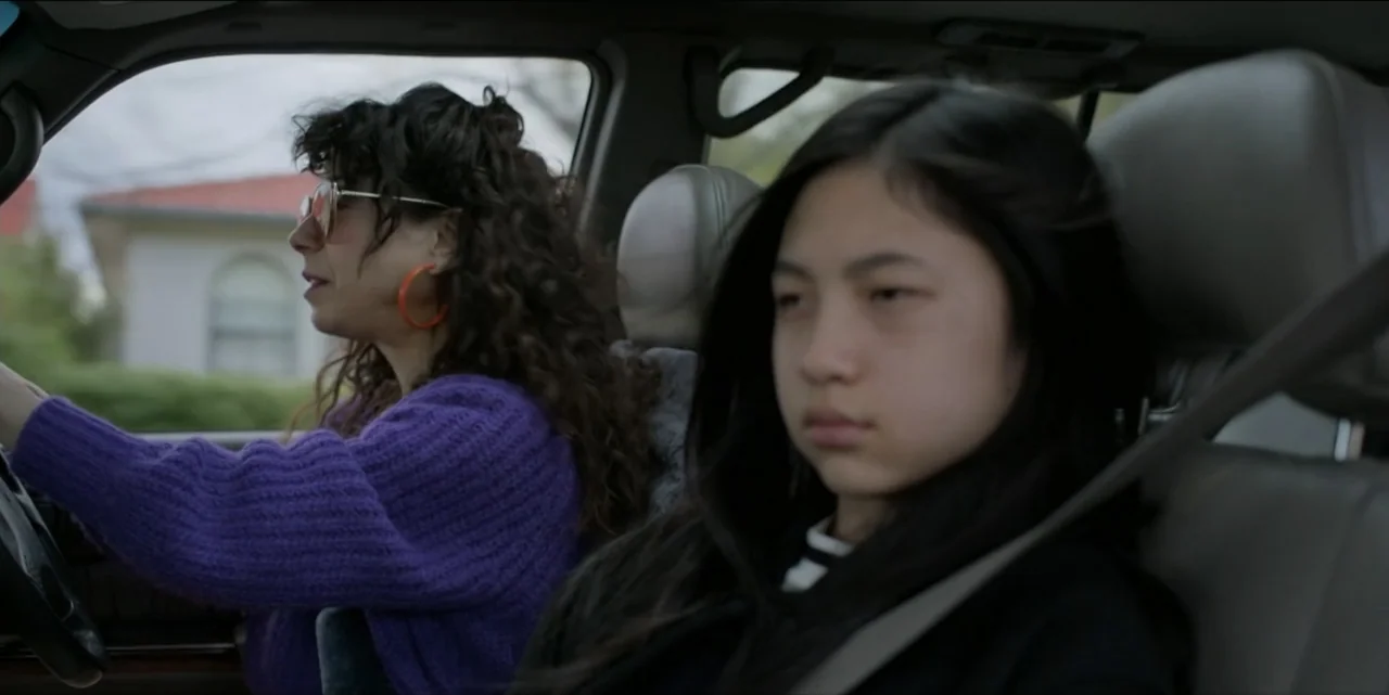 Bring Her Back (2025) movie still: medium — Two individuals sit inside the cabin of a car during the day. A woman drives while a teenage girl…