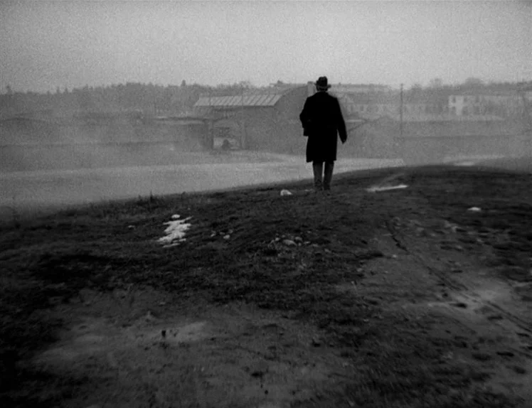 Prison (1949) movie still: wide — A man walks across a desolate, muddy field towards an industrial building in fog. The setting is a sparse,…