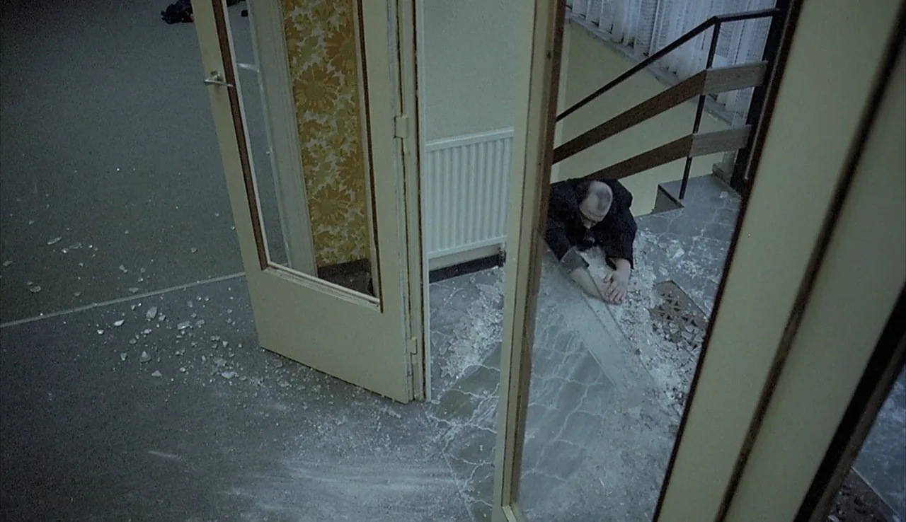Angst (1983) movie still: wide — A residential stairwell landing is covered in broken glass shards near an open doorway. A man lies prone on the…