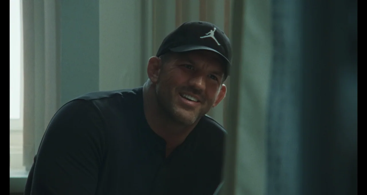 The Smashing Machine (2025) movie still: medium — A room interior features a man wearing a dark cap and shirt. The man turns his head and smiles…