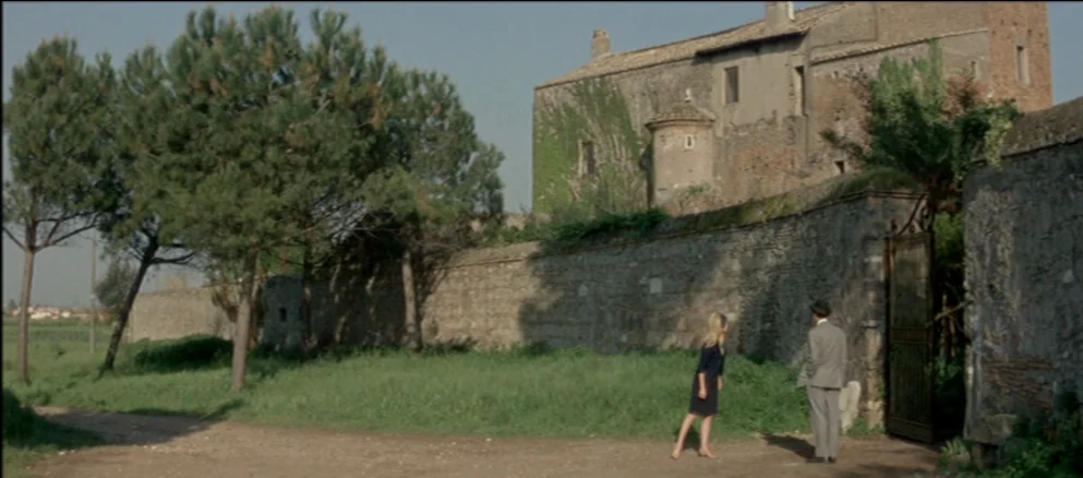 Contempt (Le Mépris) (1963) movie still: wide — An ancient stone structure covered in ivy sits behind a long perimeter wall. A woman and a man in a…