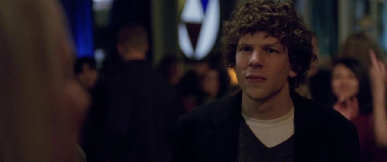 The End of the Tour (2015) movie still: medium — A crowded social gathering with dim indoor lighting and blurry party guests. A young man with curly…