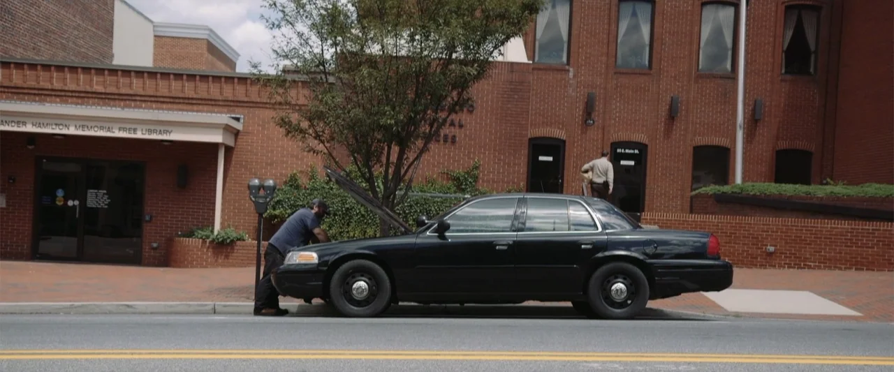 Darkness Waits (2020) movie still: wide — A brick building with signs and a sidewalk featuring a black sedan parked. A man works on the engine of a…
