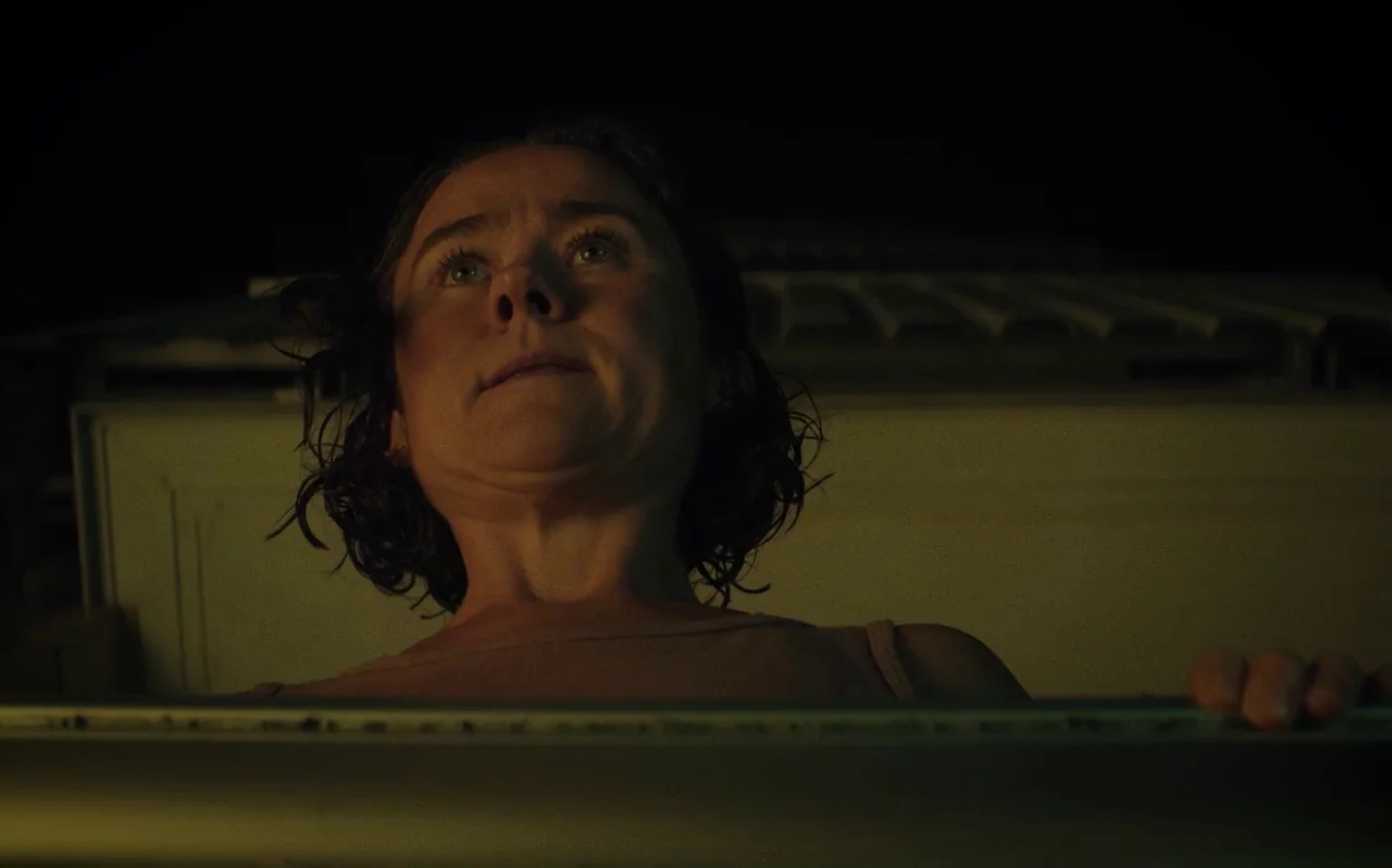 Knocking (2021) movie still: closeup — A dark rooftop environment at night with a metallic ledge in the foreground. A woman looks up from behind the…