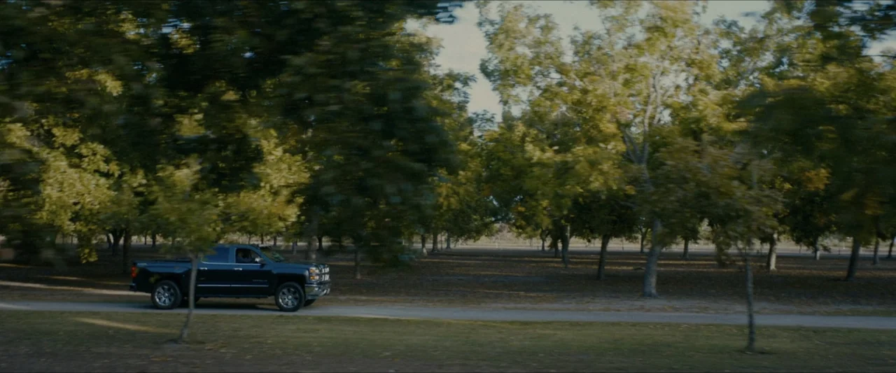 Ambush (2020) movie still: wide — A park with mature trees and a long paved path during daylight hours. A dark pickup truck drives along the road…