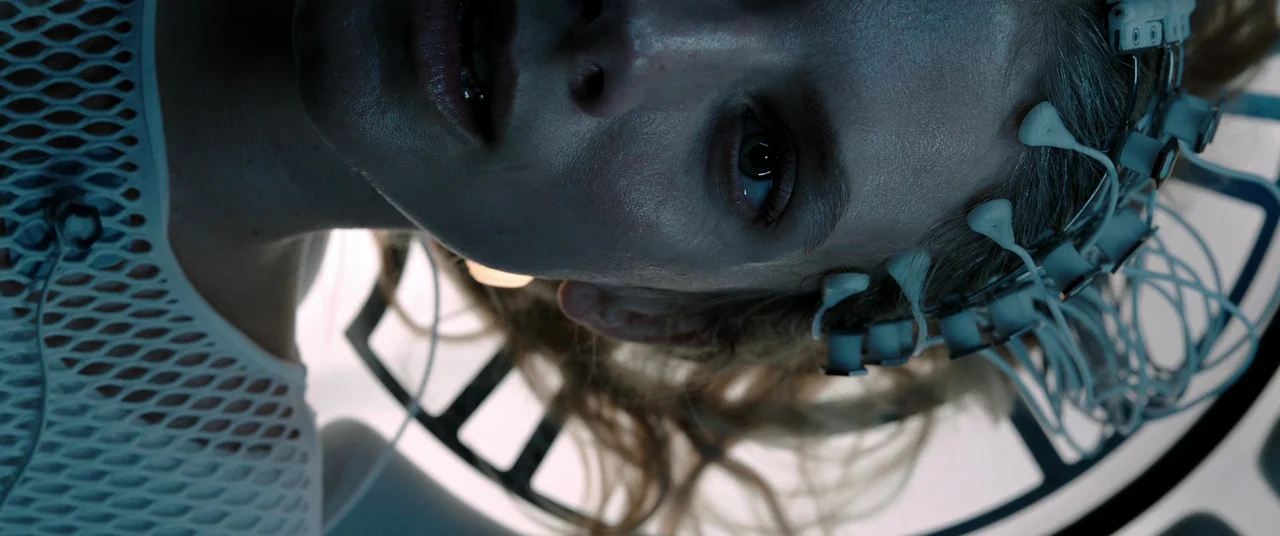 Oxygen (2021) movie still: closeup — A clinical laboratory space featuring a metallic medical device frame in the background. A woman lies with an…