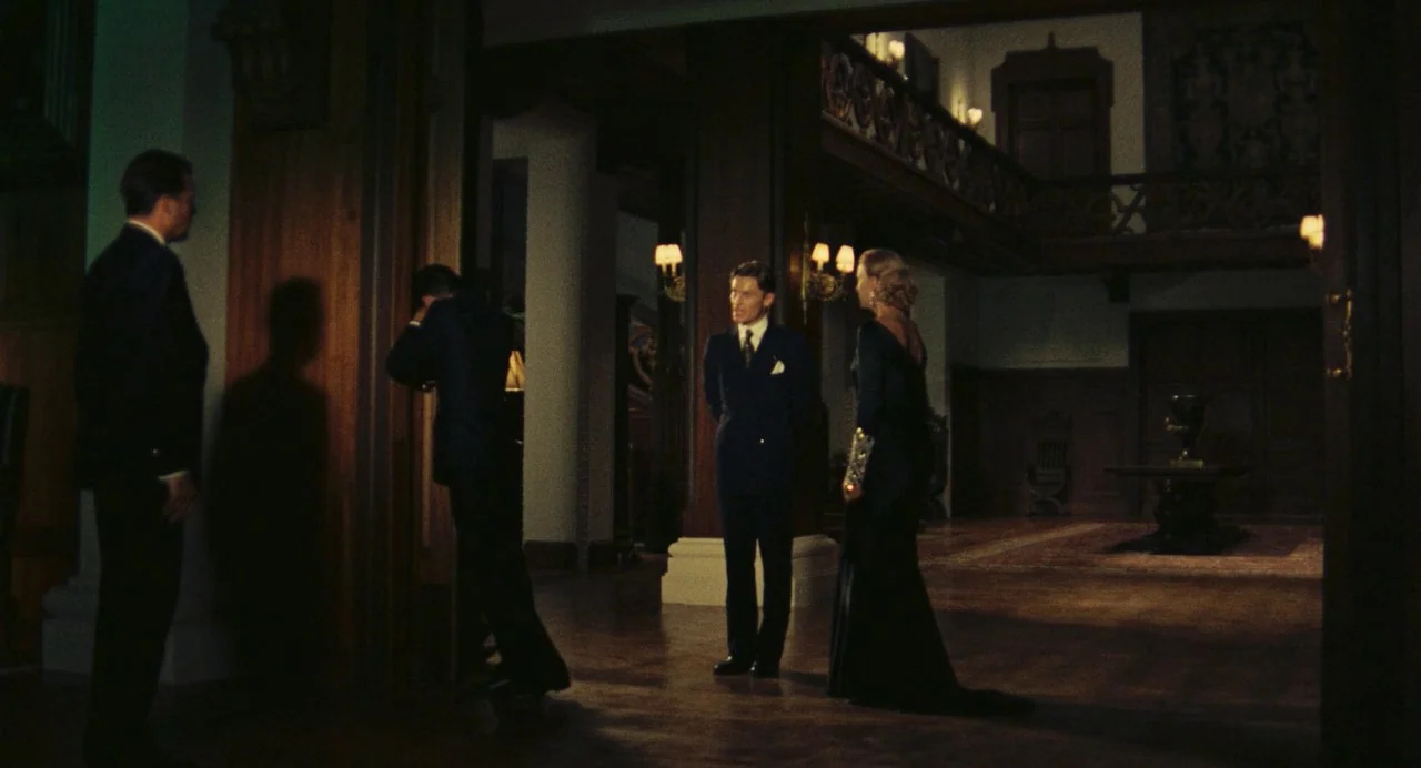 The Damned (1969) movie still: wide — A spacious mansion foyer features dark wood paneling, a staircase, and a central pillar. Four people in formal…