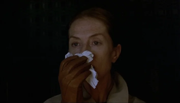 The Piano Teacher (2001) movie still: closeup — A woman sits in a dark, indistinct interior setting with minimal environmental detail. She holds a…