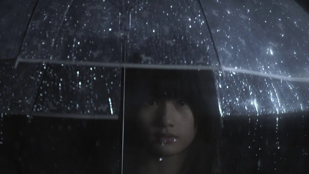 Confessions (2010) movie still: closeup — A person stands at night behind a transparent umbrella covered in rain droplets. The environment is dark…