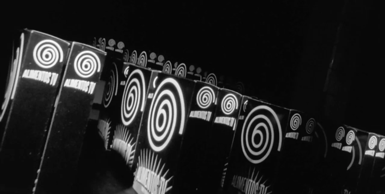 The Aerial (2007) movie still: closeup — A rows of black boxes with white spiral logos sit on a flat surface. Each box features the text Alimentos…