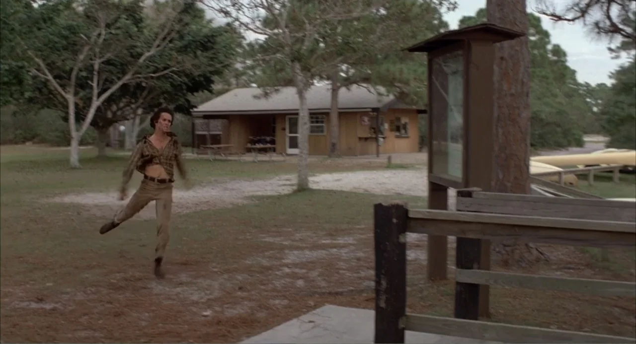 Empire of the Ants (1977) movie still: fullbody — A recreational area features a wooden cabin, a tall information board, and sparse trees. A slim…