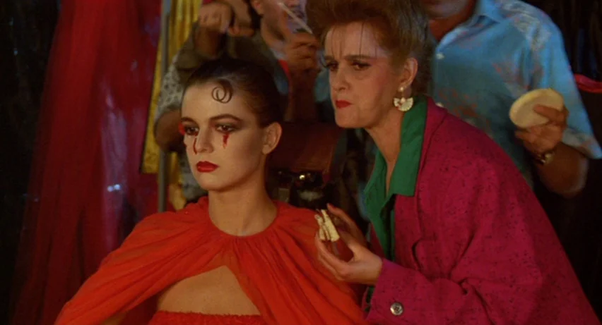 Matador (1986) movie still: medium — A crowded dressing room features rack of clothes in the background and bright artificial lights. A woman in a…
