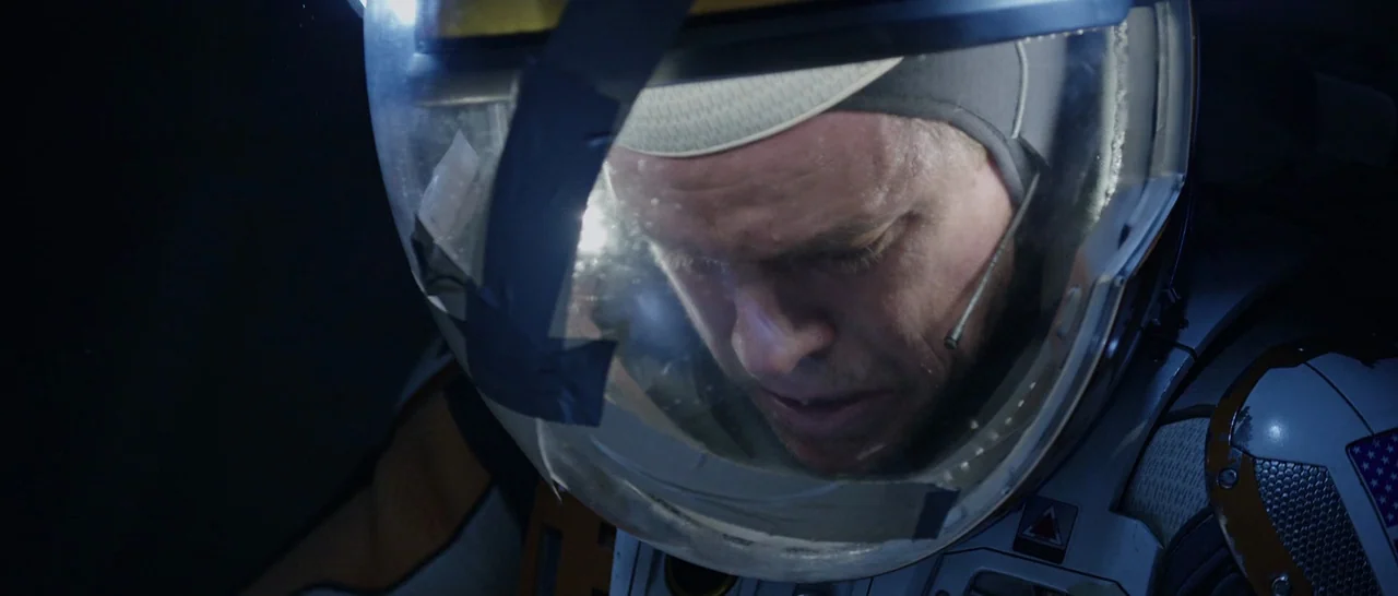The Martian (2015) movie still: closeup — A dark, cramped spacecraft cockpit interior features a man wearing a clear helmet. The subject sits still…