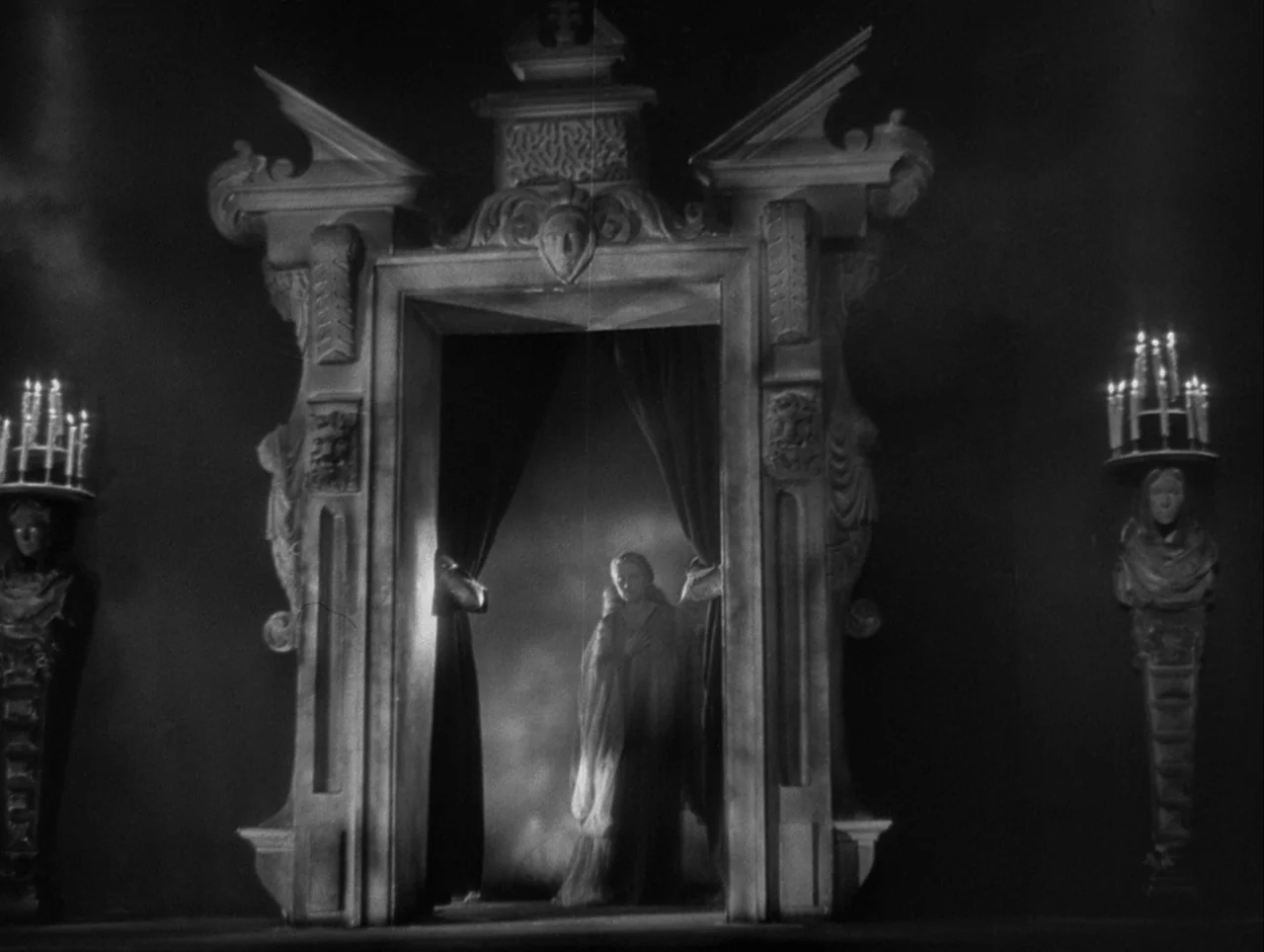 La Belle et la Bête (1946) movie still: wide — An ornate gothic stone doorway stands in a dark chamber between two candle sconces. A woman stands…