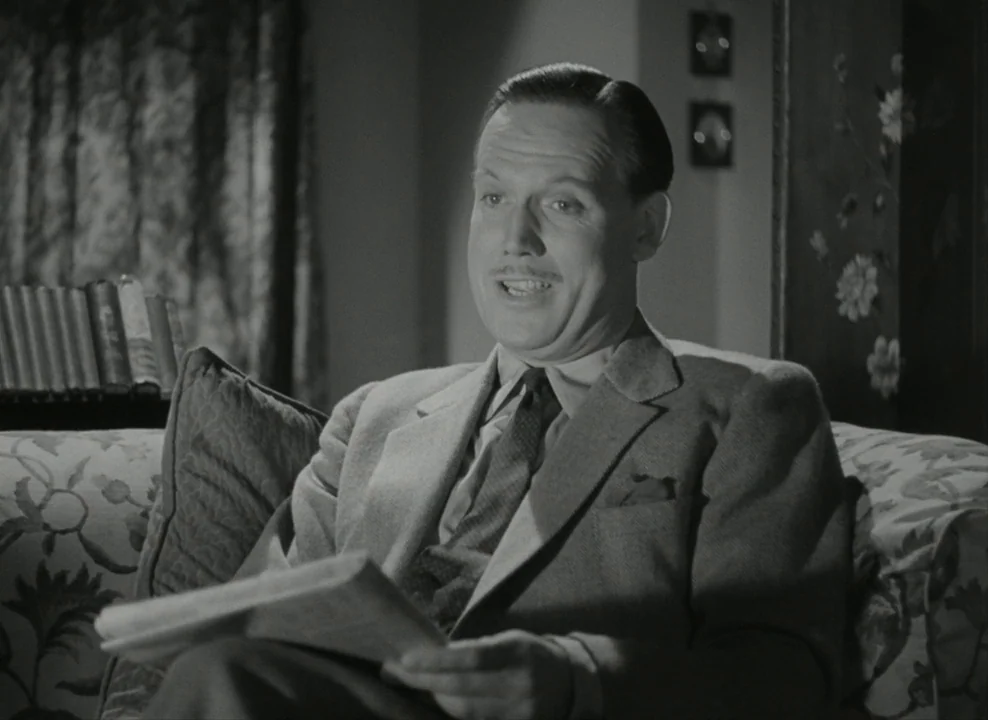 Brief Encounter (1945) movie still: medium — A well-lit interior space featuring a patterned sofa and a bookshelf in the background. A man in a suit…