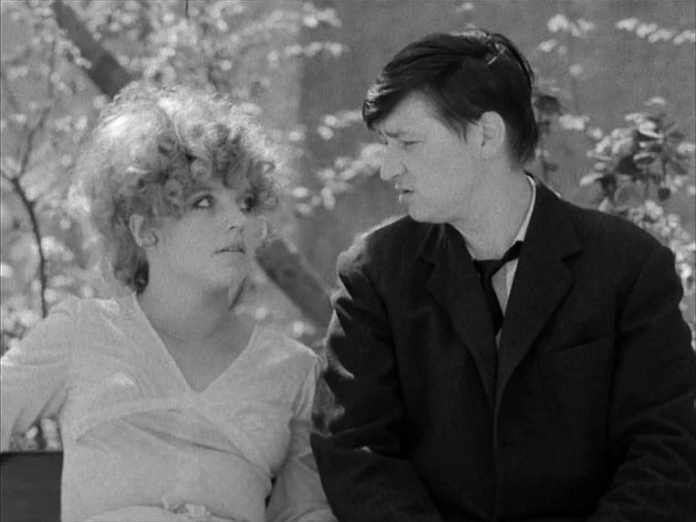 Katzelmacher (1969) movie still: medium — A man and woman sit side-by-side in a park with out-of-focus trees behind them. The two individuals are…