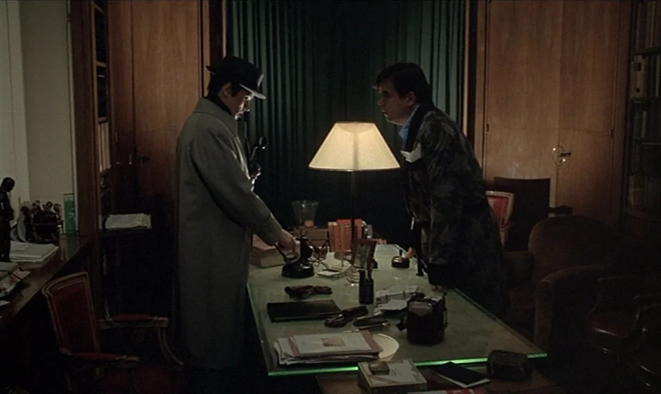 Mr. Klein (1976) movie still: medium — A wood-paneled room is dominated by a glass-topped desk and a green desk lamp. One man stands in a trench…