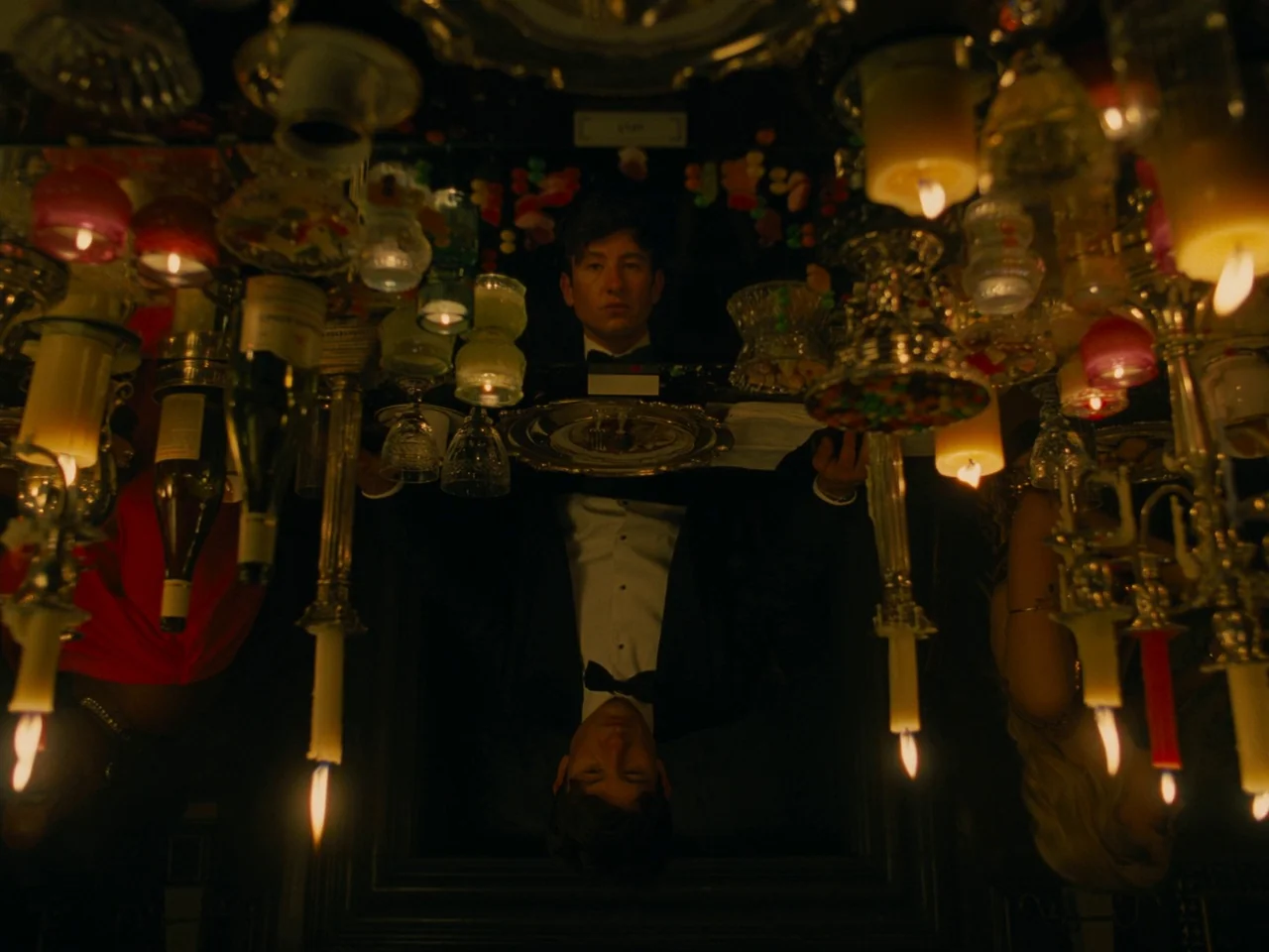 Saltburn (2023) movie still: medium — A man in a tuxedo sits at a table covered in candles and glassware. His image is reflected upside down in the…
