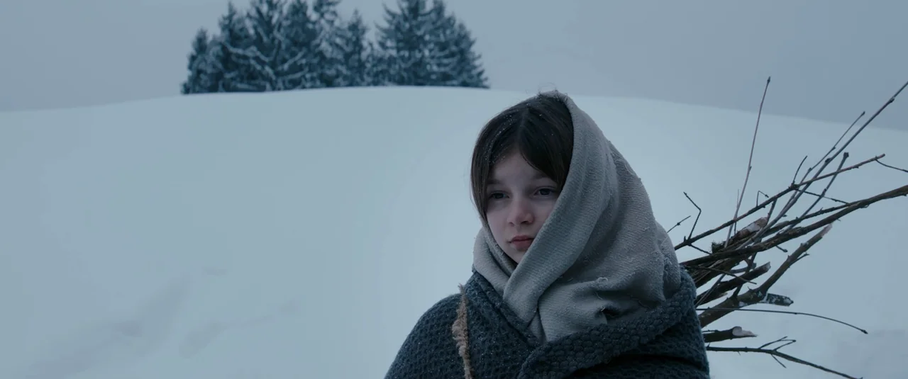 Hagazussa (2017) movie still: medium — A snow-covered hill slopes upward toward a small cluster of evergreen trees. A young girl wrapped in a heavy…