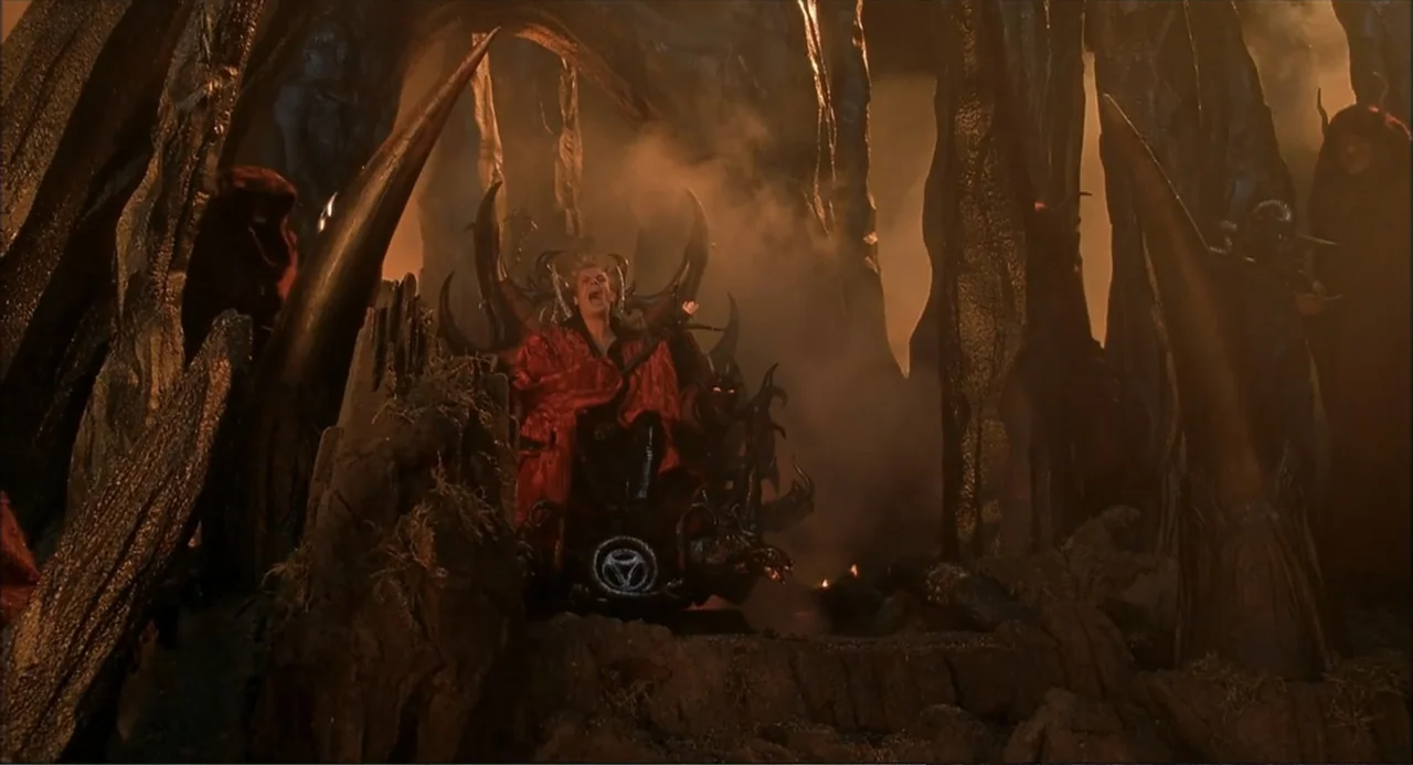 Little Nicky (2000) movie still: wide — A dark rocky cavern contains a sharp-edged throne surrounded by jagged natural stone formations. A man in a…