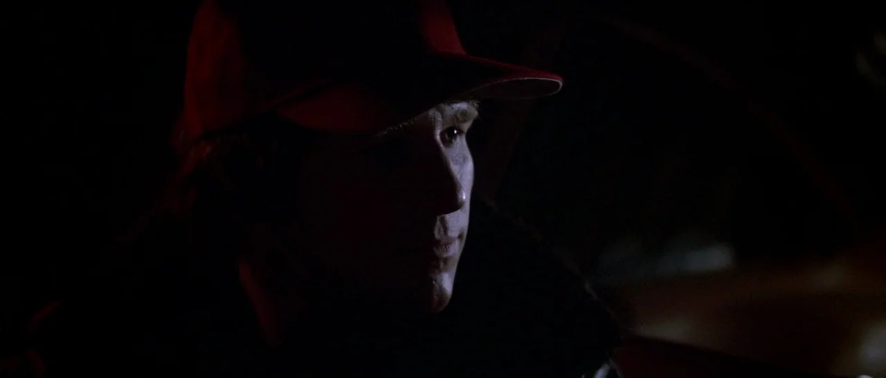 Starman (1984) movie still: closeup — The interior of a vehicle at night with darkness encompassing the background. A man wearing a baseball cap…