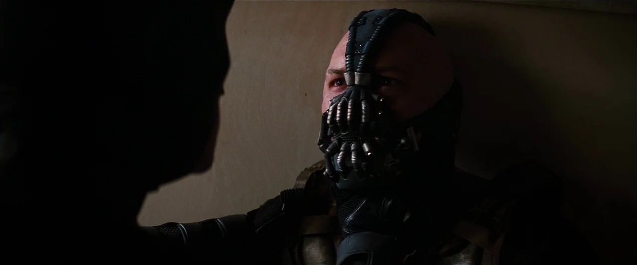 The Dark Knight Rises (2012) movie still: medium — A dimly lit interior space features a man with a large metal medical mask. The man sits against a…