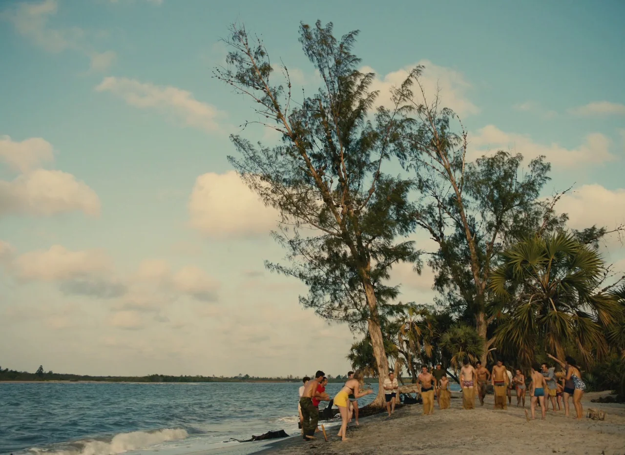 Red Island (2023) movie still: wide — A group of people competes in a sack race on a sandy coastline.