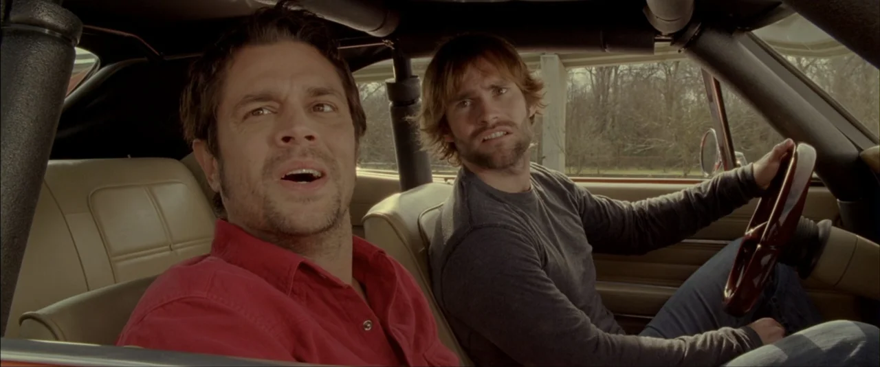 The Dukes of Hazzard (2005) movie still: medium — Two men are seated inside a car with beige leather seats. One man is driving while the other sits…