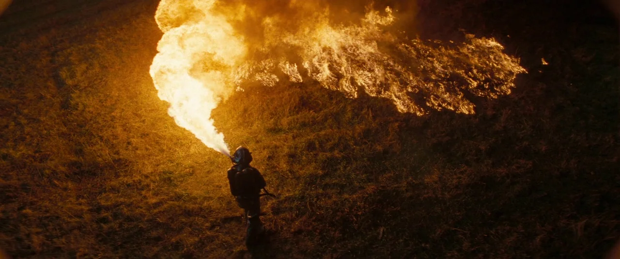 Super 8 (2011) movie still: wide — An open dark field at night is covered in dry grass and vegetation. A single individual in protective gear sprays…