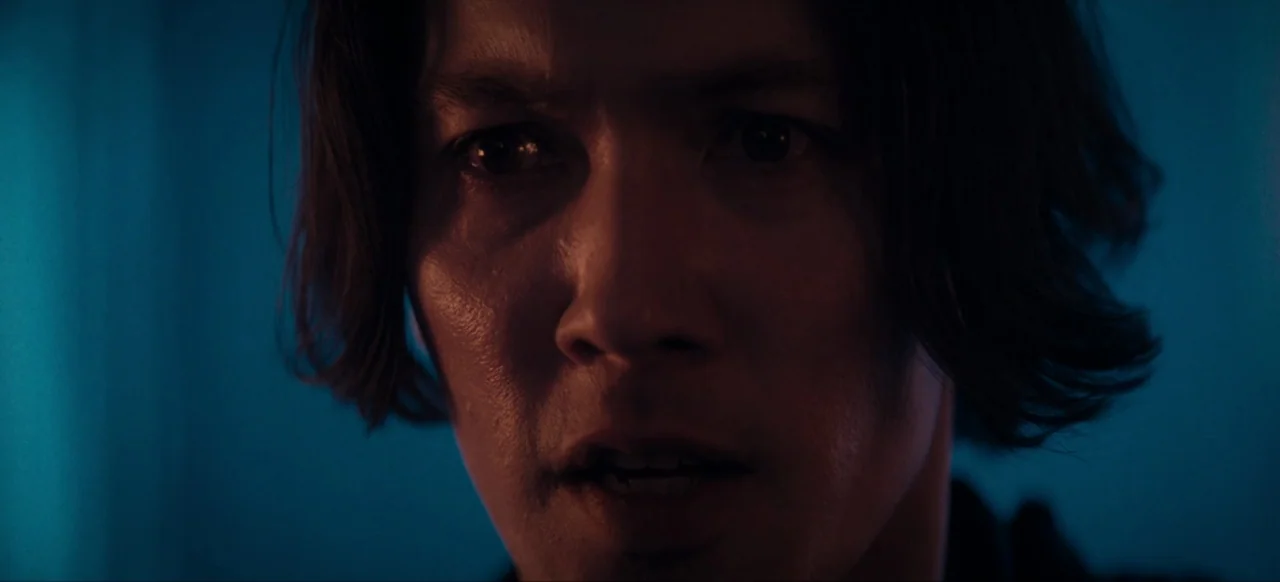 Housewife (2017) movie still: closeup — A dark space is illuminated by high-contrast red and blue light sources. A young man stares directly ahead…