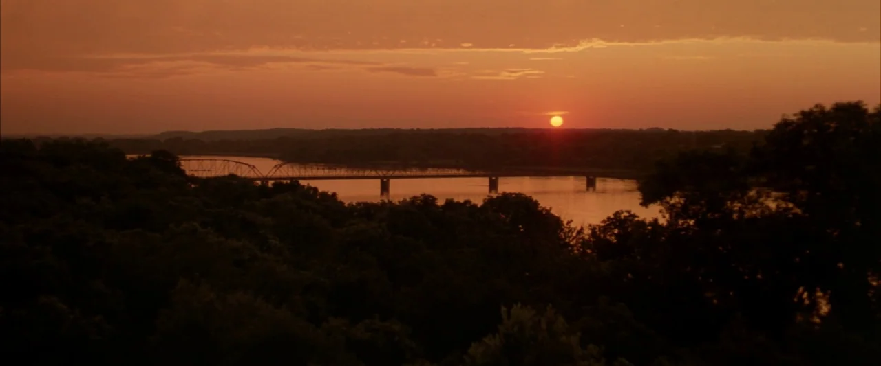 Twister (1996) movie still: establishing — A wide view of a river with a metal bridge stretching across the water. Dense trees line the riverbank…